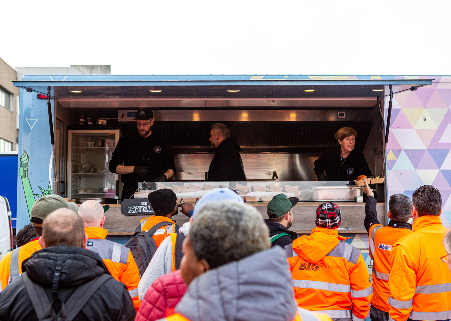 Drei Menschen verteilen Burger aus einem Foodtruck heraus an Menschen in BLG Schutzjacken (Foto)