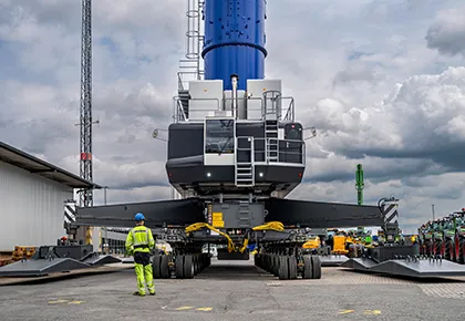 Luftaufnahme eines großen Schwerlastfahrzeugs mit vielen Rädern, das eine hohe blaue Industrieanlage transportiert; ein Arbeiter in Warnkleidung steht daneben 