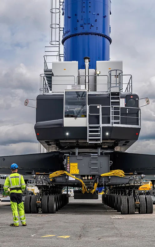 Aerial view of a large heavy transport vehicle with many wheels carrying a tall blue industrial structure; a worker in safety gear stands beside it 